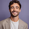 Smiling man with curly hair and beard wearing a light gray blazer over a white shirt against a purple background.
